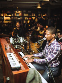 A man in a plaid shirt plays a keyboard and sings into a microphone in a crowded, dark room.