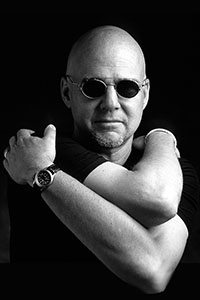 Black and white portrait of a bald man wearing round sunglasses with his arms crossed.