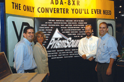 Four people posing in front of a yellow and black Prism Sound banner at an exhibition.