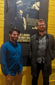 Two men pose in front of a dark movie poster for The Prodigy on a yellow brick wall.