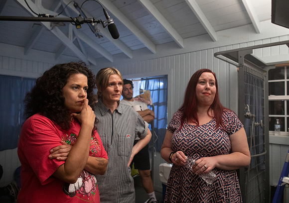 Three women stand together in a room with light blue walls and a white ceiling.