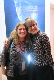 Two women smiling and embracing in front of a large blue portrait of a person.