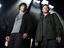 Two men standing together in dark clothing against a smoky, backlit background.