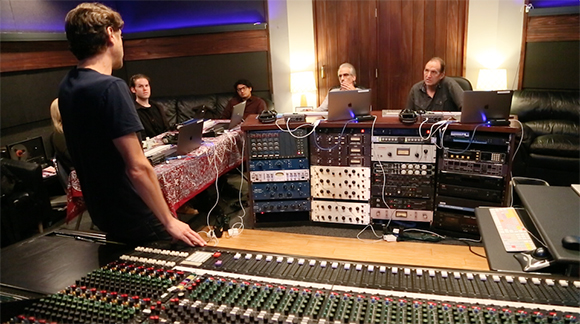 People sit in a recording studio surrounded by audio equipment and a large mixing console.