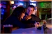 A man and woman sitting at a bar, smiling and laughing together in dim lighting.