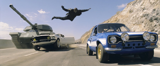 A man leaps through the air between a moving tank and a blue car on a desert road.