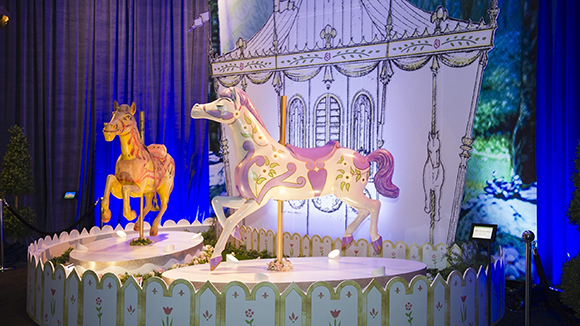 A white unicorn and a tan horse on a carousel stage against a forest backdrop.