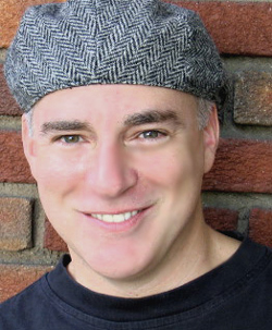 A smiling man with short hair wearing a grey herringbone flat cap against a brick wall.