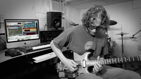 A man with curly hair playing an electric guitar in a studio with a computer, keyboard, and drum kit.