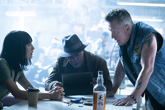 A woman and a man in a hat look at a tablet while a man in a denim vest leans over a table.