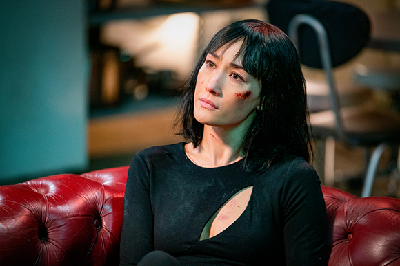 A woman with dark hair and bruises on her face sits on a red leather couch.