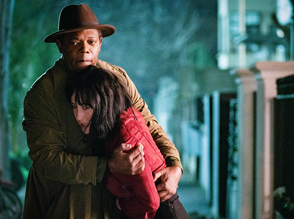 A man wearing a brown fedora holds a woman in a red jacket in a close embrace outdoors.