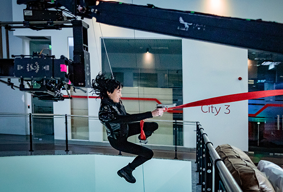 A person in black leather clothing suspended by wires in a modern building with a camera crane overhead.