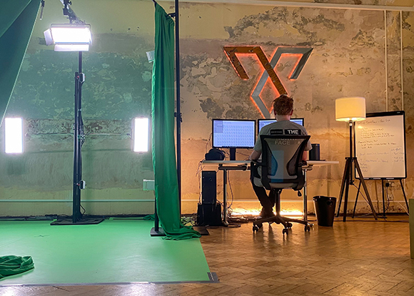 A person sits at a desk with dual monitors in a studio featuring a large glowing orange logo on a textured wall.