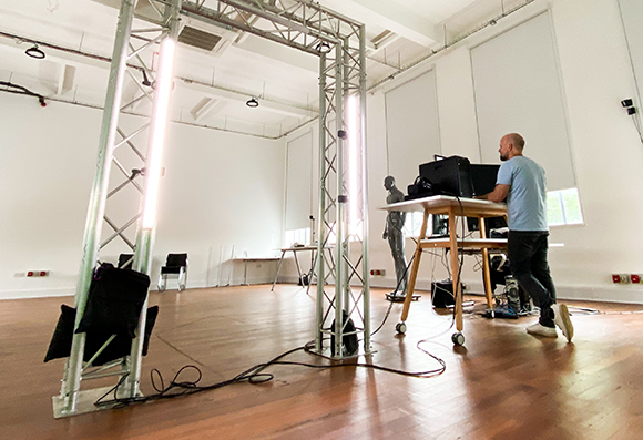 A man in a blue shirt works at a desk in a bright, large studio containing metal lighting trusses.