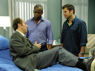 A man in a suit sits on a blue bed while two men stand over him in a room.