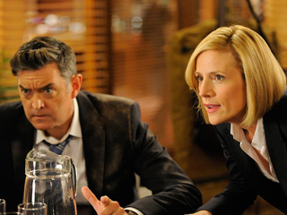 A man in a dark suit looks skeptical while a blonde woman in a dark blazer speaks intensely.