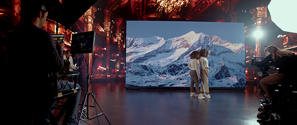 Film crew in a dark studio filming two people in front of a large screen showing snowy mountains.