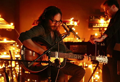 A man with long hair and glasses plays an acoustic guitar in a room filled with warm string lights.