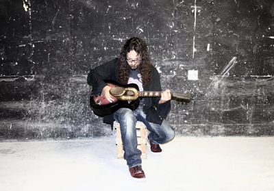 A man with long dark hair sits on a wooden crate playing an acoustic guitar against a dark, textured wall.