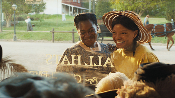 Two Black women in period clothing smile warmly in a sunlit outdoor village setting.