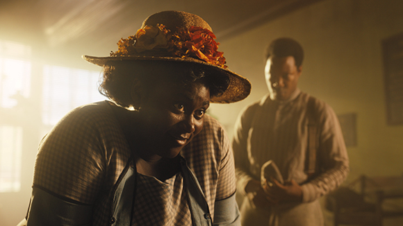 A woman wearing a straw hat with orange flowers leans forward in a sunlit, hazy room.