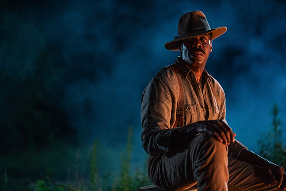 A man wearing a wide-brimmed hat and suspenders sits in a dark, smoky setting.