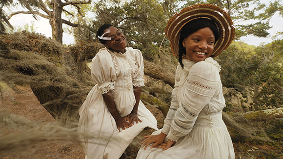 Two Black women in white period dresses smiling while sitting in a wooded, rocky landscape.