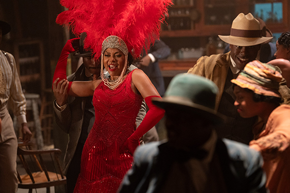 A woman in a red sequined dress and large red feather headpiece dances in a crowded room.