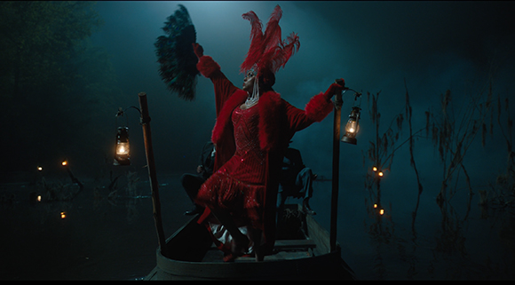 A performer in a red feathered outfit dances on a small boat illuminated by lanterns in a dark, misty swamp.