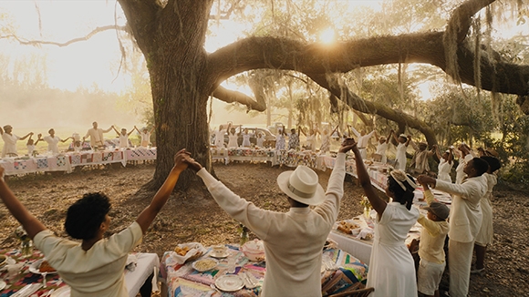People in white clothing hold hands in a circle under a large tree at sunset.