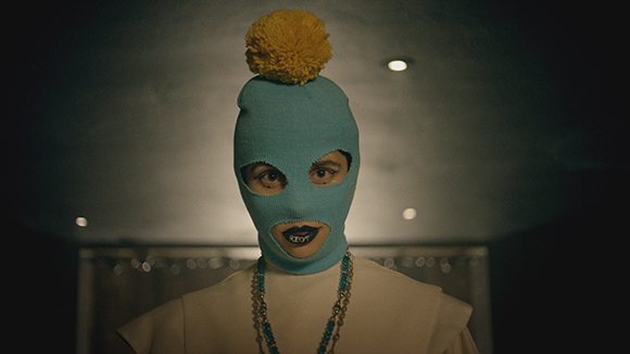 A person wearing a light blue knit balaclava with a large yellow pom-pom on top and dark lipstick.