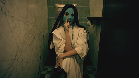 A person in a white habit and black veil wearing a blue mask, holding a finger to their lips in a restroom.