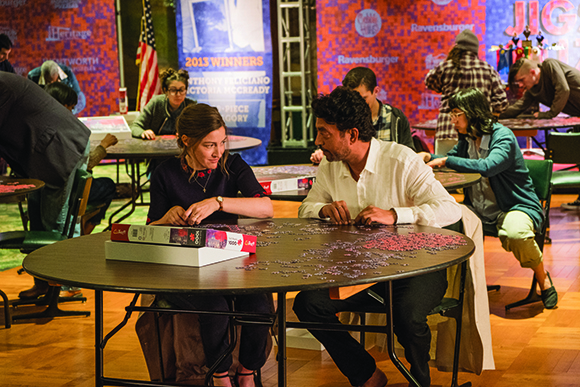Several people sit at round tables working on various jigsaw puzzles in a brightly lit room.