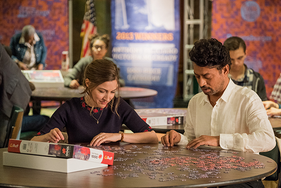 Several people sit at round tables working on large jigsaw puzzles in a room with banners.