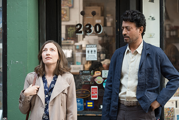 A woman in a tan trench coat looks upward while a man in a blue jacket stands beside her near a glass door.