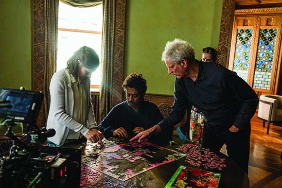 Three people lean over a dark table to assemble a large, colorful jigsaw puzzle in a sunlit room.