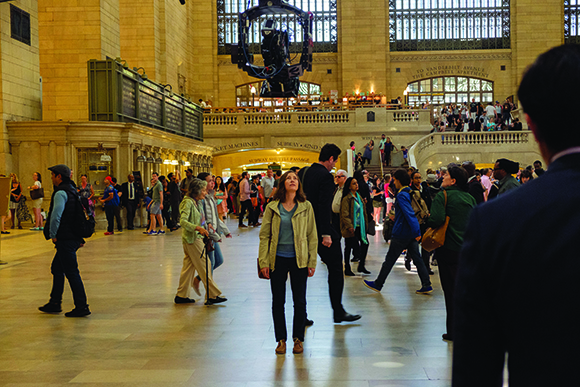 People walk through a grand, high-ceilinged train station concourse with a large mechanical device hanging above.