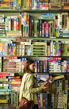A woman in a light jacket looks at a board game on a shelf filled with many colorful game boxes.