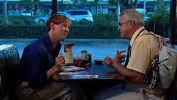 A younger man in a blue shirt and an older man in a tan shirt sit at a diner table talking.