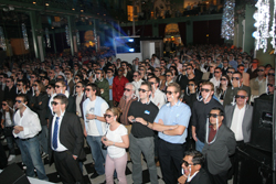 A large crowd of people standing in a dimly lit indoor venue, all wearing dark sunglasses.