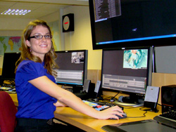 A smiling woman in a blue shirt sits at a desk with several computer monitors and a keyboard.