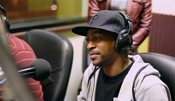 A man in a black cap and headphones sits in a chair near a microphone in a studio setting.
