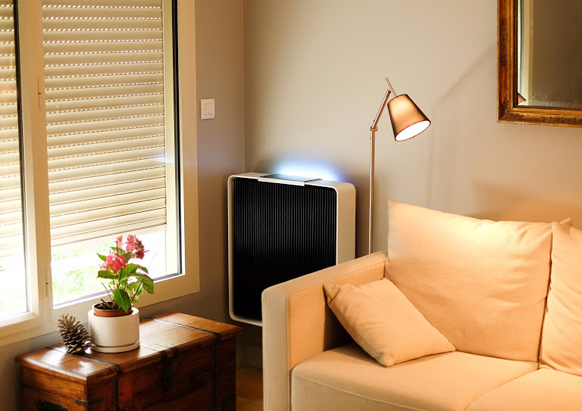 A white air purifier with a black grill stands next to a beige sofa and a window with blinds.