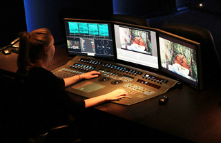 A person uses a large video editing control surface with three large monitors in a dark studio.