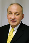 A man with light skin and thinning hair wearing a black suit, white shirt, and yellow patterned tie.