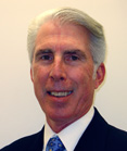 A smiling man with short gray hair wearing a dark suit, white shirt, and blue patterned tie.