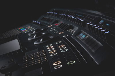 A dark, high-angle view of a complex professional audio mixing console with illuminated buttons and sliders.