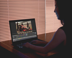 Silhouette of a person using a laptop to edit video footage in front of window blinds.
