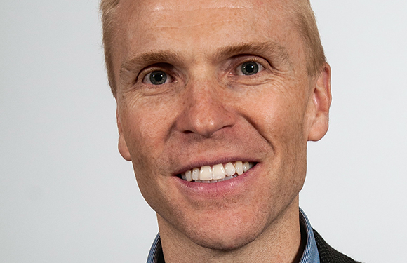 Close-up portrait of a fair-skinned man with light hair and blue eyes smiling against a white background.